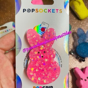 May include: Pink PopGrip phone grip with a glitter finish and a Peeps-inspired design. The grip is packaged on a white card with the PopSockets logo and the text "PopGrip".