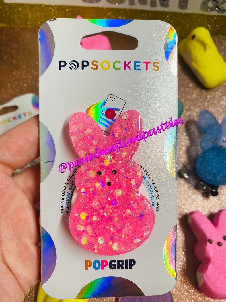 May include: Pink PopGrip phone grip with a glitter finish and a Peeps-inspired design. The grip is packaged on a white card with the PopSockets logo and the text "PopGrip".
