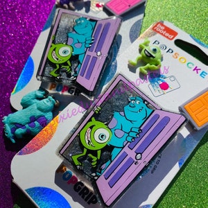 May include: A purple and silver phone grip with a glitter background featuring the characters Mike and Sully from the movie Monsters Inc. The grip has the text "PopGrip" on the bottom.