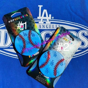 May include: Two phone grips with a baseball design. The grips are blue with red stitching and the letters "LA" in white. The grips are on packaging that says "PopSockets", "PufGrip", "Game of Thrones", and "#1" and "#2".