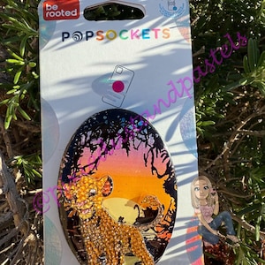 May include: A PopSockets phone grip featuring a sparkling Simba design from The Lion King. The grip has a black background with a sunset scene and the Simba character. The PopSockets packaging is white with the brand name and logo.