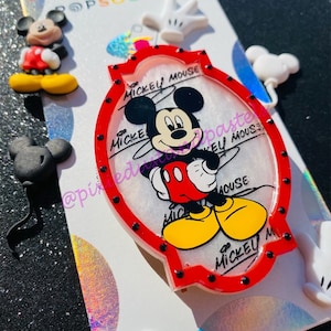 Può includere: Uno sfondo bianco con un PopSocket di Mickey Mouse rosso e bianco. Il PopSocket presenta un'immagine di cartone animato di Topolino con le parole "Mickey Mouse" scritte intorno all'immagine. Ci sono diversi altri PopSockets a tema Topolino sullo sfondo.
