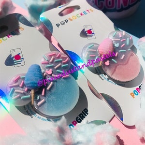 May include: Two pink and blue phone grips shaped like Mickey Mouse ears with cotton candy and sprinkles on top. The grips are on packaging that says "PopSockets" and "PopGrip".
