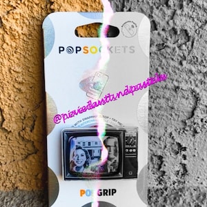 Könnte beinhalten: Ein weißer PopSockets-Handygriff mit einem Schwarzweißbild eines Vintage-Fernsehers. Der Fernsehbildschirm zeigt eine Frau und einen Mann. Der Text "POPGRIP" ist auf der Unterseite des Griffs aufgedruckt.