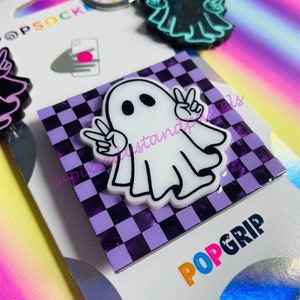 May include: A white ghost with peace signs on a purple and black checkered background. The ghost is a phone grip with the word "POPGRIP" printed below it.