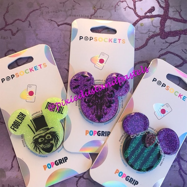 May include: Three phone grips with glitter designs. The first grip is green with a black and white image of a ghost. The second grip is purple with a black image of a ghost. The third grip is purple with a green and white striped design.