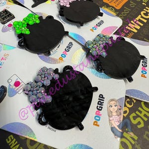 May include: Black cauldron PopSockets with decorative crystal accents in pink, green, and iridescent colors. The PopSockets are on a white and iridescent background. The word "POPGRIP" is visible.