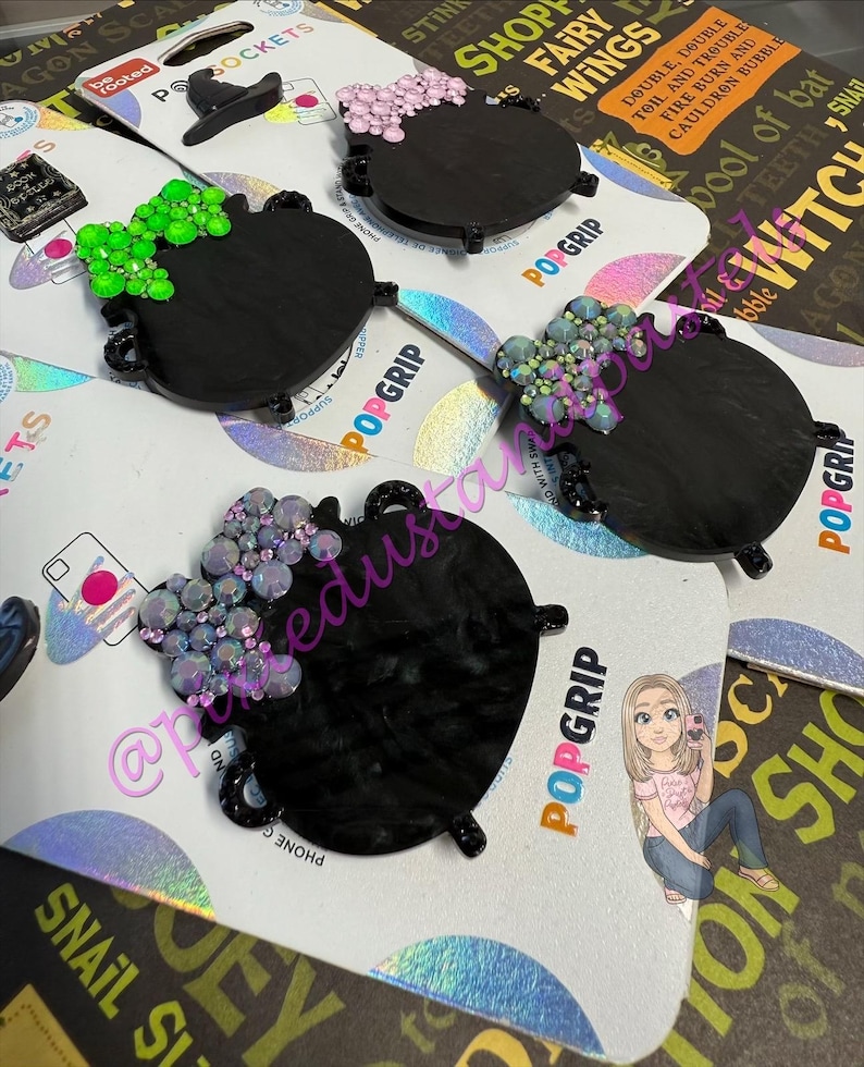 May include: Black cauldron PopSockets with decorative crystal accents in pink, green, and iridescent colors. The PopSockets are on a white and iridescent background. The word "POPGRIP" is visible.