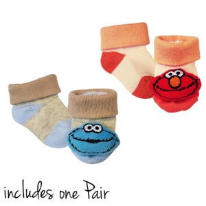 May include: A pair of baby socks with character appliques. One pair features a blue Cookie Monster applique and the other pair features a red Elmo applique. The socks are beige with blue and red accents. "includes one pair" is written below the socks.
