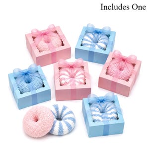 May include: Pink and blue donut-shaped washcloths, some in gift boxes with ribbons. The boxes are either pink or blue, with a clear window to see the washcloth. The text "Includes One" is visible.
