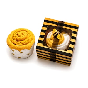 May include: Two decorative towels, one in a silver container with bee accents and the other in a black and gold striped box. The towels are rolled to resemble a flower. The box is adorned with a bee-shaped embellishment and a black ribbon.