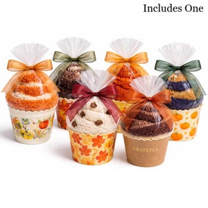 May include: Six decorative cupcake-shaped gifts, each with a different colored towel inside. The towels are wrapped in clear cellophane and tied with a ribbon. The cupcake cups have fall-themed designs, including pumpkins and leaves. One cup says "Grateful".