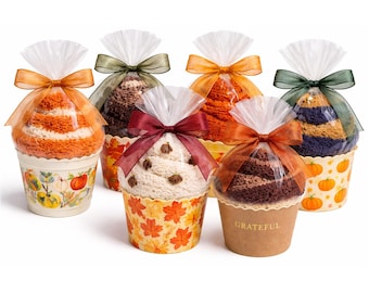 May include: Six decorative cupcake-shaped gifts, each with a different colored towel inside. The towels are wrapped in clear cellophane and tied with a ribbon. The cupcake cups have fall-themed designs, including pumpkins and leaves. One cup says "Grateful".