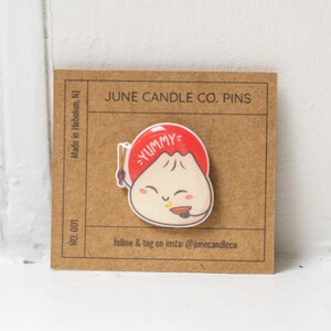 May include: A white and red enamel pin featuring a cartoon dumpling with a red hat that says "YUMMY" and chopsticks. The pin is on a brown card with the text "JUNE CANDLE CO. PINS" and "follow & tag on insta: @junecandleco".