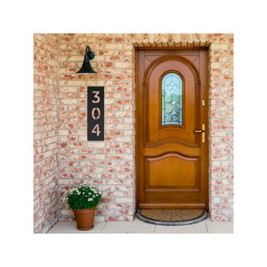 May include: A black metal house number sign with the number 304 displayed on a brick wall. The sign is mounted next to a wooden door with a glass window.