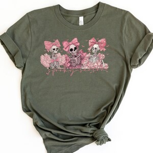 May include: Olive green t-shirt featuring a graphic of three skeletons wearing pink bows and tutus. The skeletons are surrounded by pink flowers, with the text "Spooky Season" in a pink script below the image. The shirt is made of a soft material.