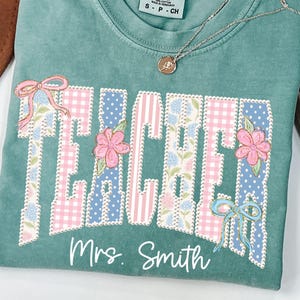 May include: A teal Comfort Colors t-shirt with the word "TEACHER" in a patchwork design featuring pink gingham, blue floral patterns, and pink flowers. The name "Mrs. Smith" is printed below the word "TEACHER". A silver necklace with a pendant is draped on the shirt.