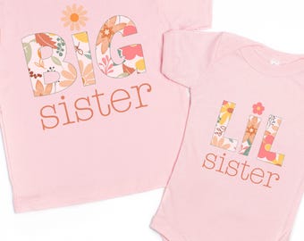 May include: Two pink shirts, one a t-shirt and the other a onesie, with the words "BIG sister" and "LIL sister" in floral print. The t-shirt has a daisy graphic above the text.