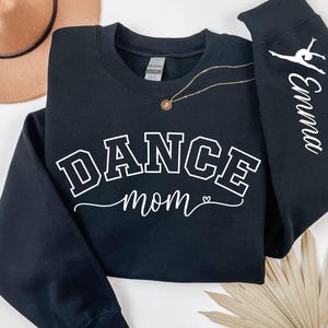 May include: Black sweatshirt with the words "DANCE mom" in white block letters. The sleeve on the right has the name "Emma" in white script with a gymnast silhouette. A gold necklace and a tan hat are in the background.