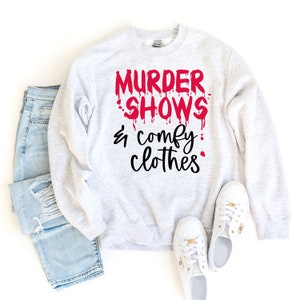 Puede incluir: Sudadera gris claro con el texto "MURDER SHOWS & comfy clothes" en rojo y negro. La imagen incluye vaqueros azul claro y zapatillas blancas.