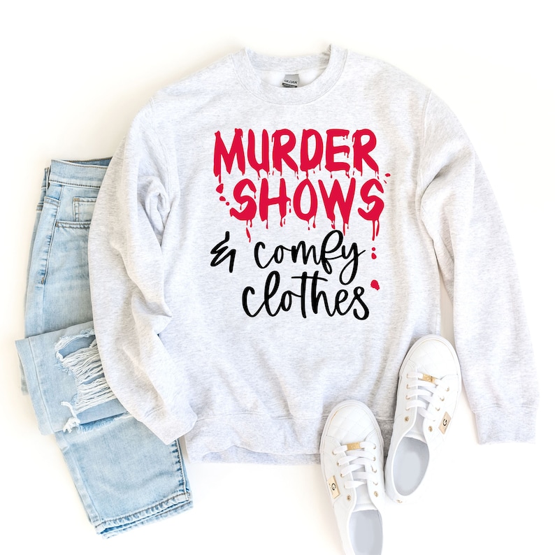 Puede incluir: Sudadera gris claro con el texto "MURDER SHOWS & comfy clothes" en rojo y negro. La imagen incluye vaqueros azul claro y zapatillas blancas.