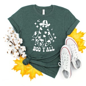 May include: A dark green t-shirt featuring a white graphic of a ghost wearing a cowboy hat with a cow print design. The text "BOO Y'ALL" is below the ghost. White sneakers and fall leaves are in the image.