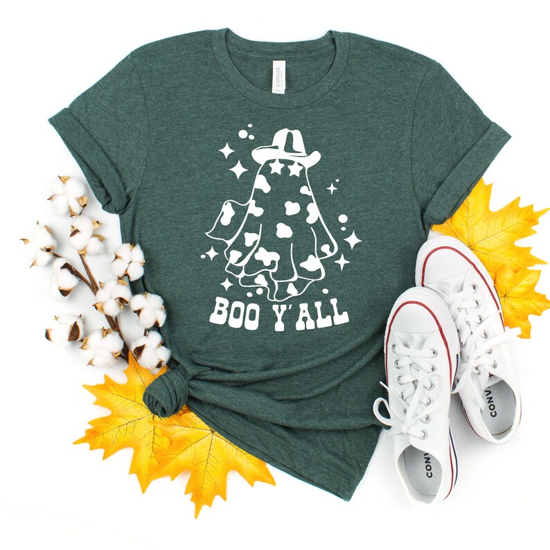 May include: A dark green t-shirt featuring a white graphic of a ghost wearing a cowboy hat with a cow print design. The text "BOO Y'ALL" is below the ghost. White sneakers and fall leaves are in the image.