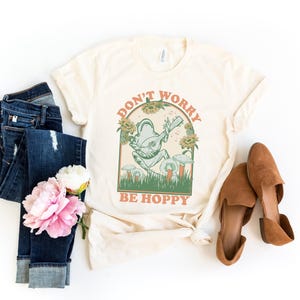 May include: A cream colored t-shirt with a green frog playing a guitar. The frog is surrounded by sunflowers and mushrooms. The text "Don't Worry Be Hoppy" is printed on the shirt.