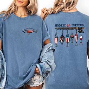 May include: A light blue t-shirt with the phrase "Hooked on Freedom" and a fishing lure design featuring the American flag. The back of the shirt displays a fishing rod with several lures, also in patriotic colors. The year "1776" is printed on the back.