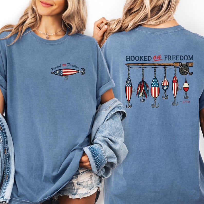 May include: A light blue t-shirt with the phrase "Hooked on Freedom" and a fishing lure design featuring the American flag. The back of the shirt displays a fishing rod with several lures, also in patriotic colors. The year "1776" is printed on the back.