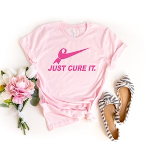 May include: Pink t-shirt with a pink ribbon and a swoosh design. The text "JUST CURE IT." is printed on the shirt.