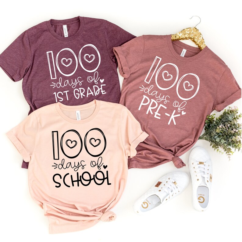 Op de afbeelding: Drie roze en kastanjebruine T-shirts met witte tekst die "100 dagen school", "100 dagen 1e klas" en "100 dagen kleuterklas" zegt.