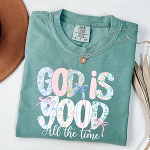 Może przedstawiać: Zielona koszulka Comfort Colors z napisem "GOD IS GOOD All the time" w patchworkowym wzorze. Litery wypełnione są kwiatowymi wzorami i pastelowymi kolorami. Złoty naszyjnik spoczywa na koszulce.