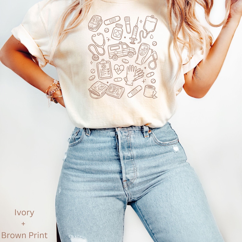 May include: Ivory-colored t-shirt with a brown print featuring medical-themed illustrations. The design includes a stethoscope, ambulance, heart monitor, and other medical tools. The shirt is paired with light blue jeans.