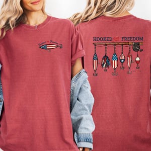 May include: A red t-shirt with the text "Hooked on Freedom" and a fishing lure design. The front of the shirt has a small lure graphic. The back features a fishing rod with several lure designs.
