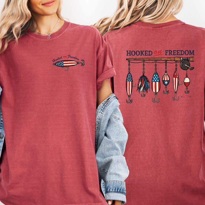May include: A red t-shirt with the text "Hooked on Freedom" and a fishing lure design. The front of the shirt has a small lure graphic. The back features a fishing rod with several lure designs.