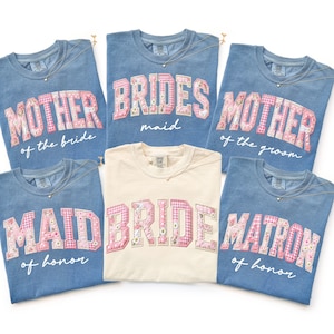 May include: A collection of blue and cream-colored t-shirts with text in a floral pattern. The shirts say "Mother of the Bride", "Bridesmaid", "Mother of the Groom", "Maid of Honor", "Bride", and "Matron of Honor". Each shirt has a small necklace.