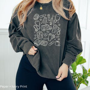 May include: Dark gray sweatshirt with a white medical-themed design. The design includes illustrations of medical tools such as a stethoscope, ambulance, and blood pressure monitor. The text "Peper + Ivory Print" is at the bottom.