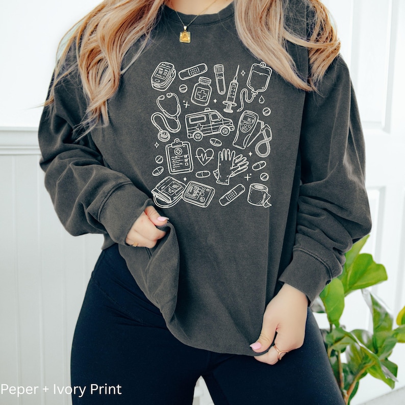 May include: Dark gray sweatshirt with a white medical-themed design. The design includes illustrations of medical tools such as a stethoscope, ambulance, and blood pressure monitor. The text "Peper + Ivory Print" is at the bottom.