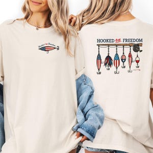 May include: Cream-colored t-shirt with the text "Hooked on Freedom" and a fishing lure graphic on the front. The back features a fishing rod with several lures decorated with the American flag and the number "1776".