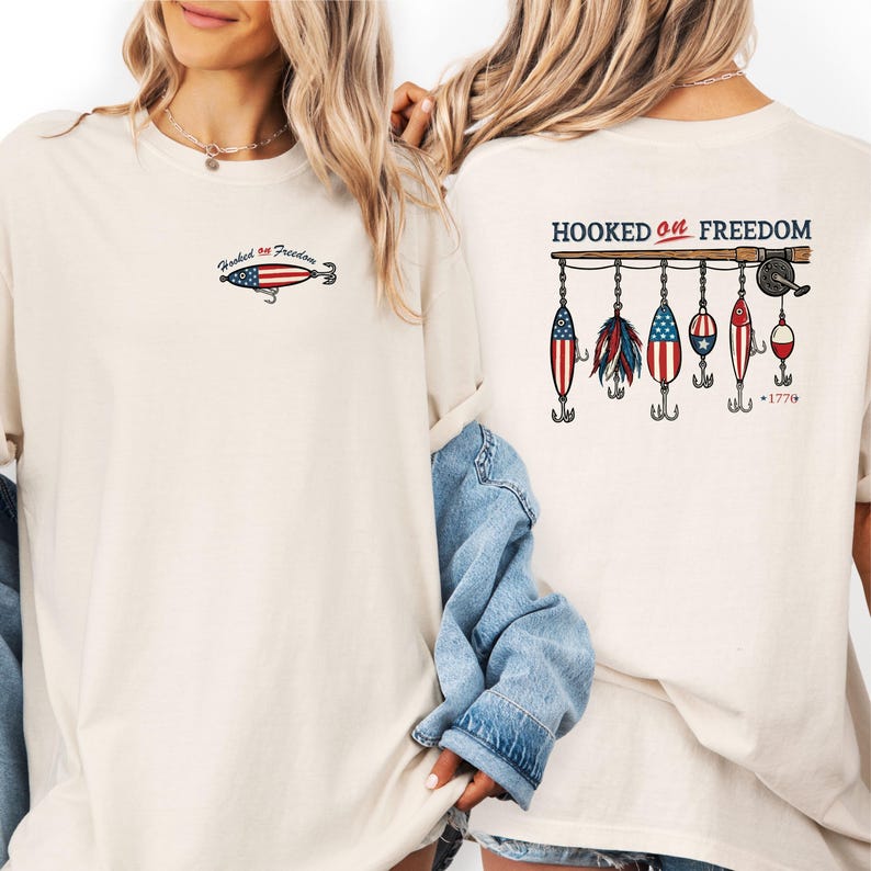 May include: Cream-colored t-shirt with the text "Hooked on Freedom" and a fishing lure graphic on the front. The back features a fishing rod with several lures decorated with the American flag and the number "1776".