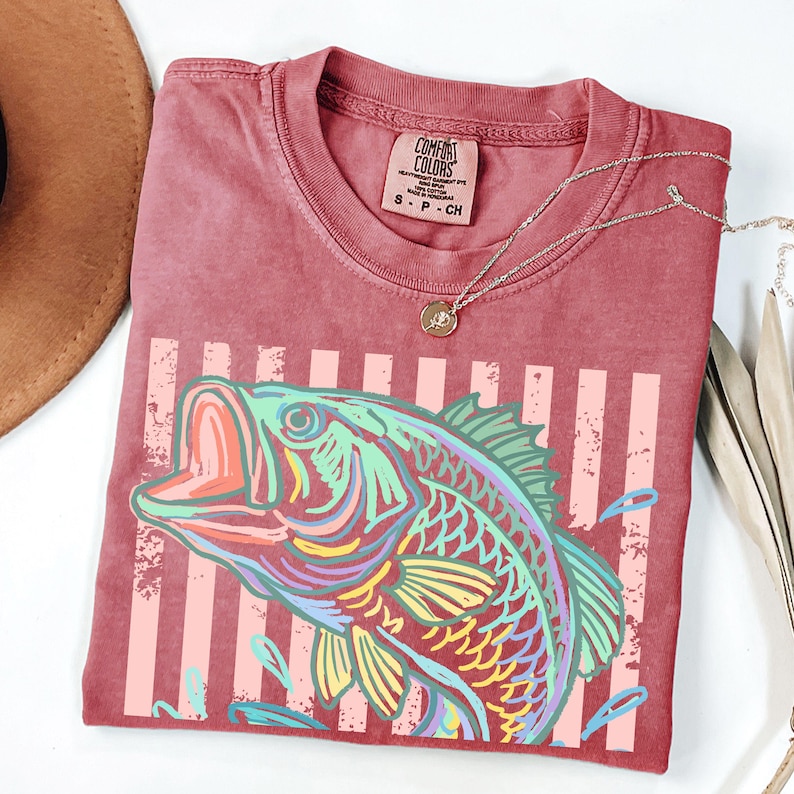 Pu&ograve; includere: Una maglietta Comfort Colors bordeaux con una grafica colorata di un pesce e un design a righe. La maglietta ha un girocollo e maniche corte. Una collana d'argento con un ciondolo &egrave; drappeggiata sulla maglietta.