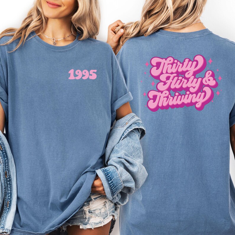 May include: A blue t-shirt with the year "1995" printed in pink on the front. The back of the shirt features the phrase "Thirty, Flirty & Thriving" in a pink, retro font with star accents. The shirt has short sleeves and a relaxed fit.
