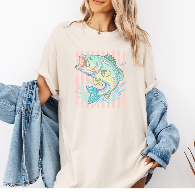 Pu&ograve; includere: T-shirt color crema con un'illustrazione colorata di un pesce che salta. Il pesce &egrave; raffigurato in tonalit&agrave; di blu, verde, giallo e rosa, su uno sfondo a righe rosa e bianche. La maglietta ha una vestibilit&agrave; ampia.