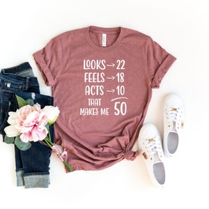 May include: A mauve-colored t-shirt with the text "LOOKS -> 22 FEELS -> 18 ACTS -> 10 THAT MAKES ME 50" in white. The shirt is styled with blue jeans, white sneakers, and a bouquet of pink and white flowers.