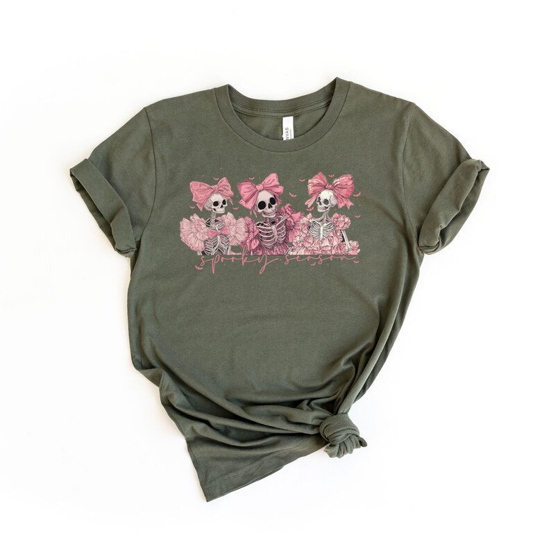 May include: Olive green t-shirt featuring a graphic of three skeletons wearing pink bows and tutus. The skeletons are surrounded by pink flowers, with the text "Spooky Season" in a pink script below the image. The shirt is made of a soft material.