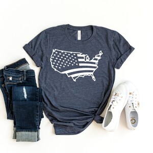 May include: A navy blue t-shirt featuring a white outline of the United States with a flag design. The shirt is paired with blue jeans and white sneakers.