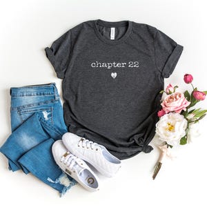 May include: A dark gray t-shirt with the text "chapter 22" and a small heart graphic. The shirt is paired with blue jeans and white sneakers. A bouquet of pink and white flowers is also present.