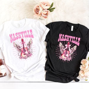 May include: Two t-shirts featuring a pink and white graphic of a guitar with wings and the word "NASHVILLE" in pink lettering. One t-shirt is white and the other is black.