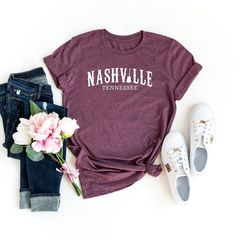 Pu&ograve; includere: Una maglietta color granata con la scritta "Nashville Tennessee" in lettere bianche. La maglietta &egrave; stesa su uno sfondo bianco con un paio di scarpe da ginnastica bianche, un mazzo di fiori rosa e un paio di jeans blu.